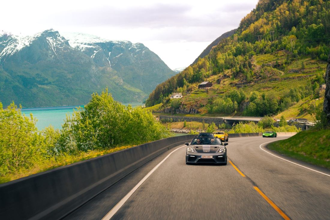 Grijze Porsche Spyder voorop een colonne supercars langs een turquoise fjord