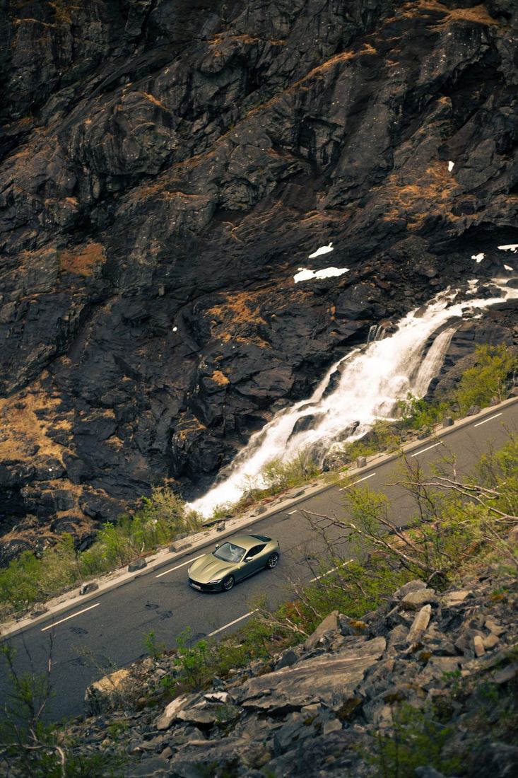 Groene Ferrari Roma op een bergweg naast een waterval, gezien van boven