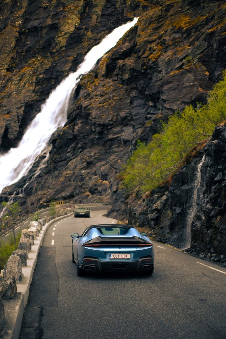 Blauwe Ferrari Roma Spider van achteren op een weg langs een waterval