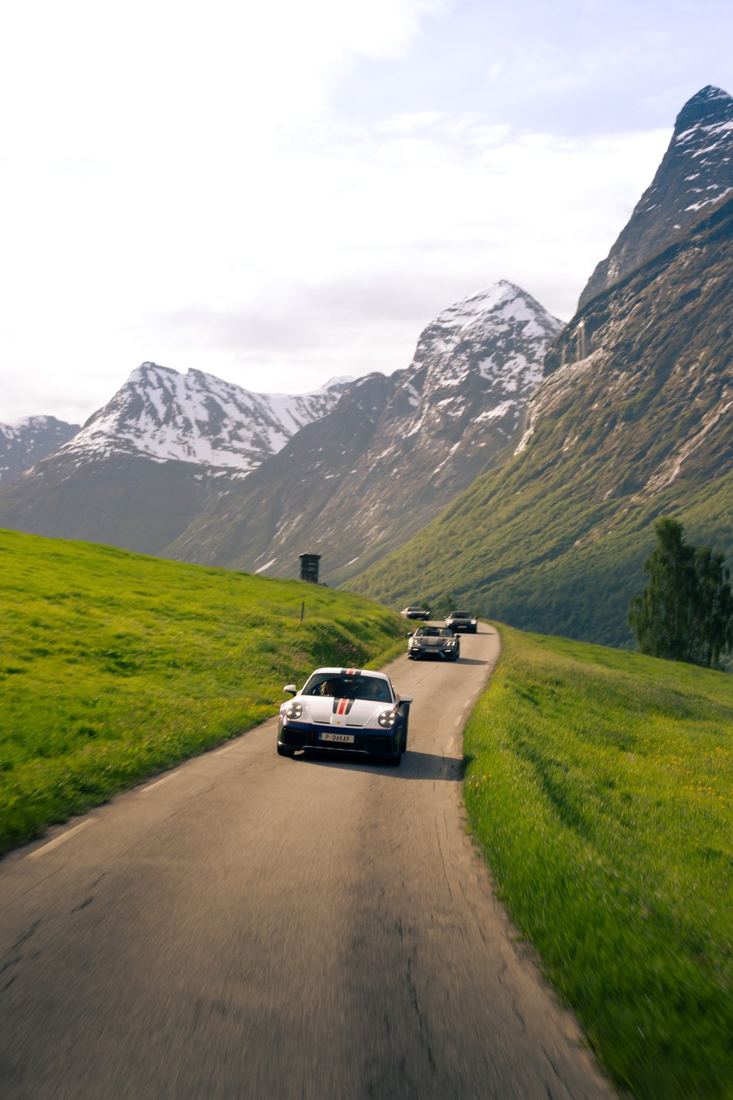 Witte Porsche 911 Dakar voorop een groep in een groene vallei met besneeuwde toppen